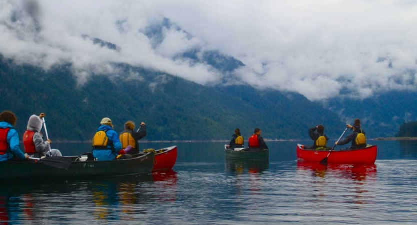 a group of outward bound students paddle canoes on calm water with foggy mountains in the background